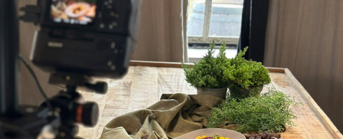 Kamera fotografiert Teller mit dekorativ angerichtetem Essen auf Tisch neben Kräutern, durch Fenster fällt Tageslicht.