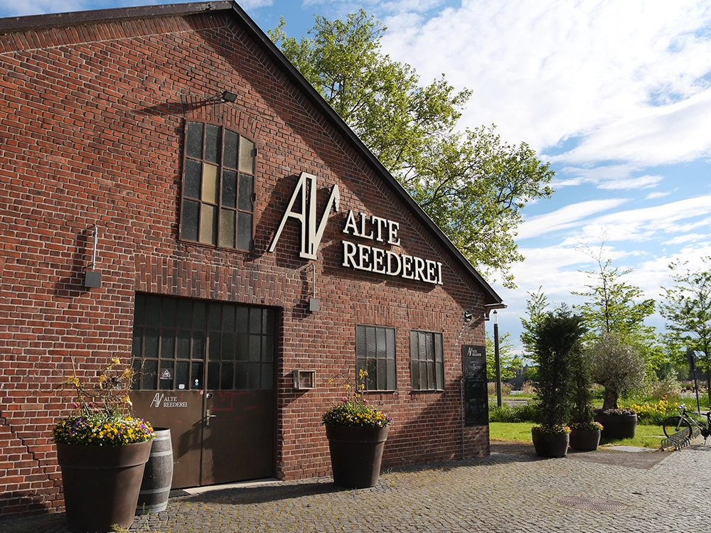 AlteReederei-Aussenansicht Backsteingebäude mit Schild Alte Reederei, umliegende Blumen und Bäume, sonniger Himmel im Hintergrund.