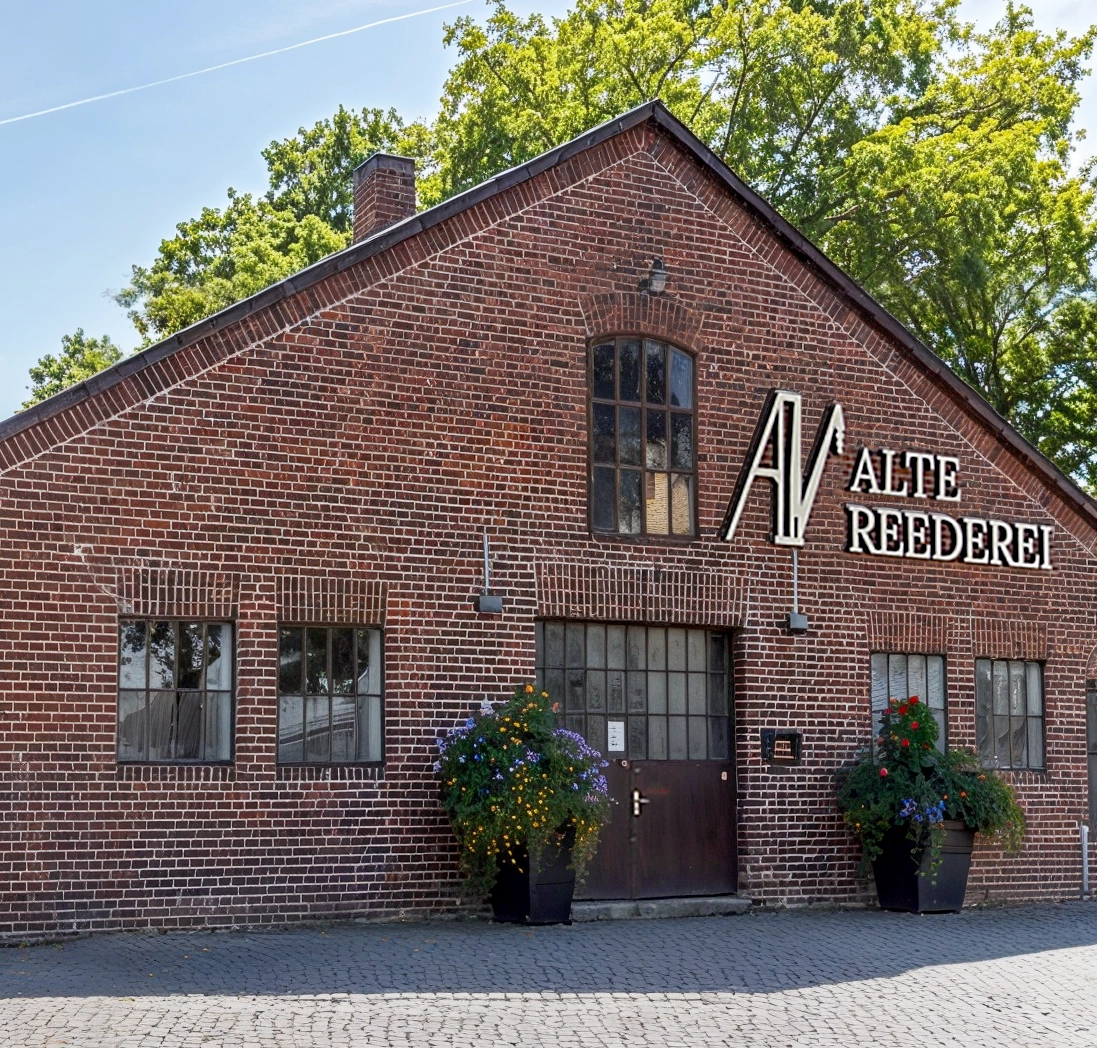 Ziegelgebäude mit Schild Alte Reederei, Pflanzen links und rechts der Eingangstür, umgeben von Bäumen.