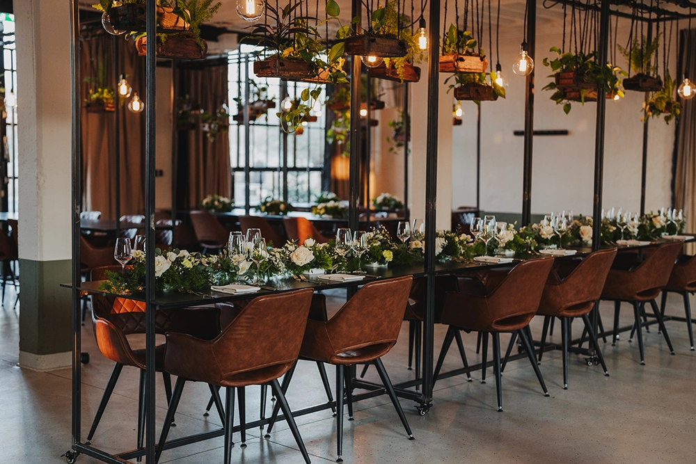 AlteReederei-Innenansicht2 Ein festlich gedeckter Tisch mit Blumen und hängenden Pflanzen in einem modernen, gemütlich beleuchteten Raum.