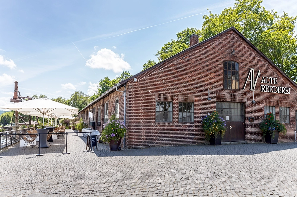 altereederei Backsteingebäude mit Aufschrift Alte Reederei, Terrasse mit Tischen und Schirmen, sonniger Tag, Bäume im Hintergrund.