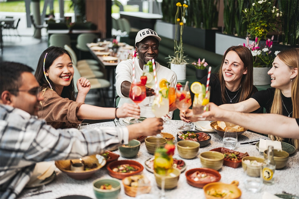 CampusGarden-Gaeste Gruppe von fünf Personen stößt im Restaurant mit Cocktails an, umgeben von Speisen auf einem Tisch, fröhliche Stimmung.