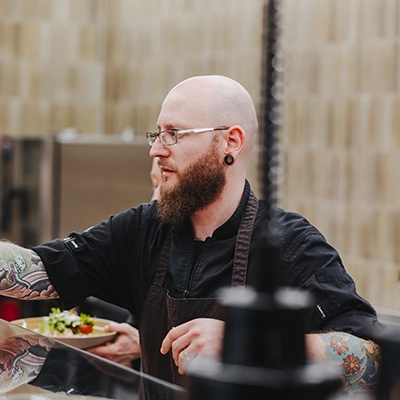 CampusGarden-Koch Ein Koch mit Glatze und Bart richtet Essen auf Tellern an, vor einer Küchenwand.