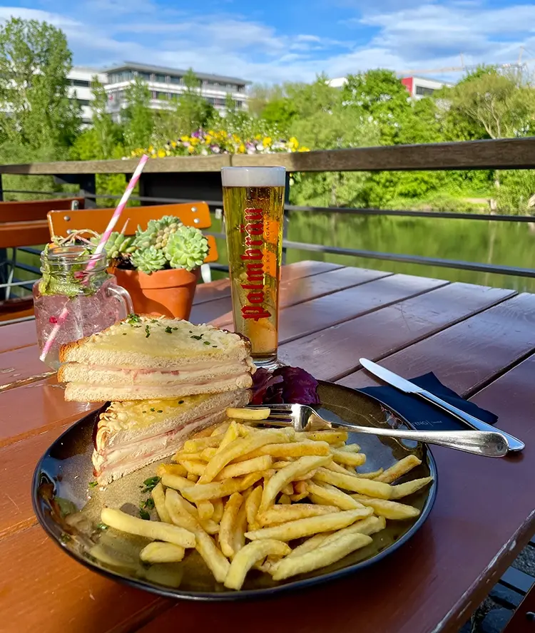Neckargarten-Gericht Sandwich und Pommes auf einem Tisch, im Freien mit Blick auf Bäume und einen Fluss, neben einem Bierglas auf der Terrasse.