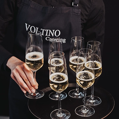 VOLTINO-Bedienung2 Eine Person in Voltino Catering-Schürze hält ein Tablett mit sechs gefüllten Champagnergläsern.