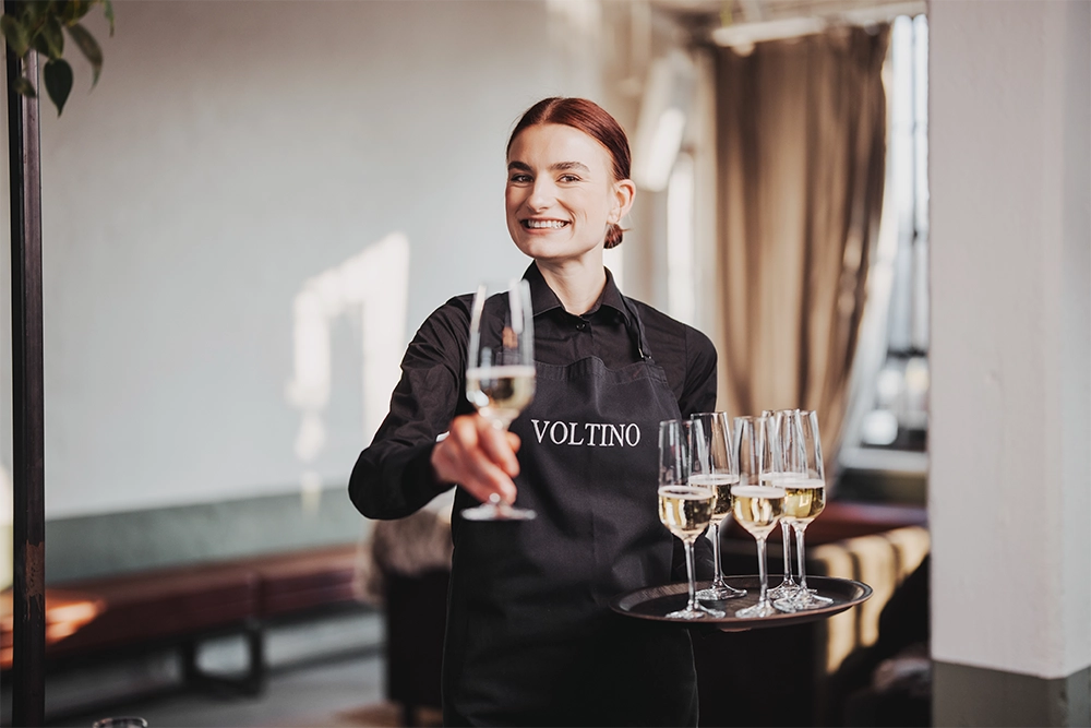 VOLTINO-Bedienung4 Eine Kellnerin in schwarzer Uniform serviert lächelnd Champagnergläser auf einem Tablett in einem hellen Raum.