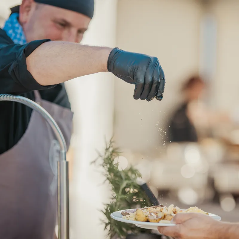 Voltino-Catering-Saltbar Koch mit blauer Haube und Handschuh würzt Gericht auf Teller in Restaurant.