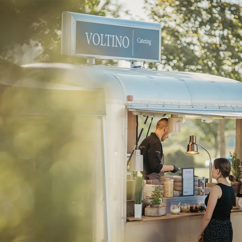 Koch bedient Kundin an einem Catering-Foodtruck im Freien, umgeben von Bäumen, mit Voltino Schild.