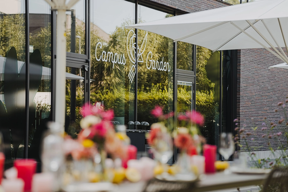 Campus Garden Außenansicht Außenbereich eines Restaurants mit der Aufschrift Campus Garden, gedeckter Tisch mit Blumen und Kerzen im Vordergrund.