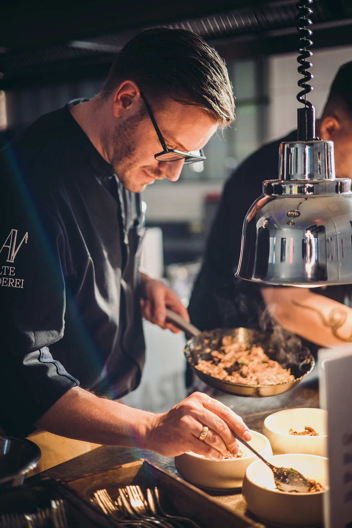 Ein Koch in schwarzer Uniform füllt dampfendes Essen in Schalen in einer Küche, beleuchtet von einer Lampe.