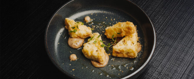Frittierte Käsestücke mit Kräutern und Sauce auf schwarzem Teller, auf dunkler Tischoberfläche präsentiert.