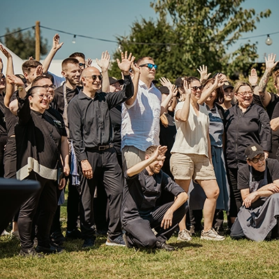 Voltino-12 Eine Gruppe von Menschen steht im Freien auf einer Wiese, alle lächeln und winken bei sonnigem Wetter.