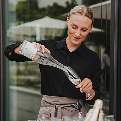 VOLTINO-Bedienung1 Frau in schwarzer Bluse schenkt Sekt in ein Glas ein, draußen bei einer Veranstaltung.