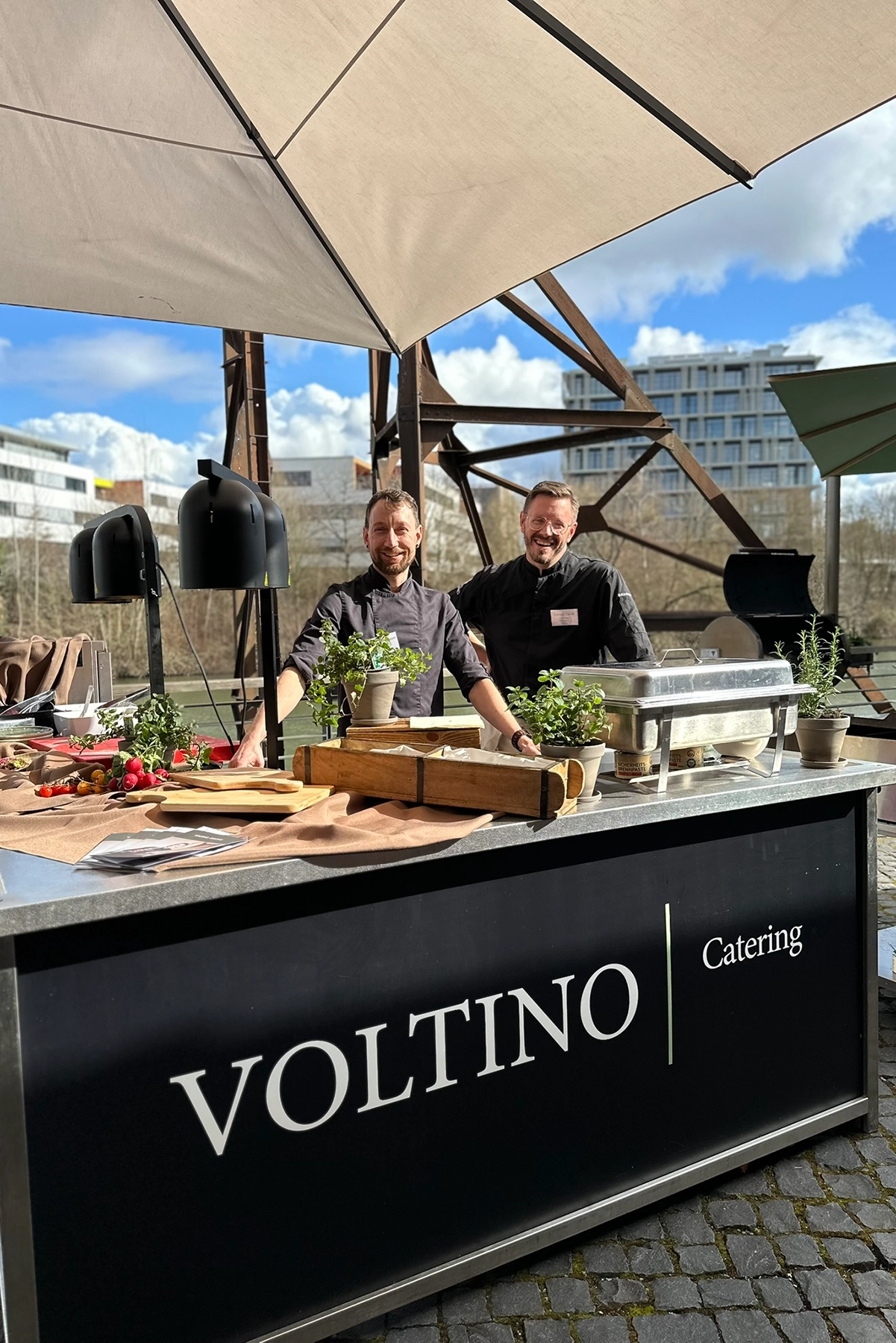 Zwei Köche stehen hinter einem Außencatering-Stand mit Töpfen, Zutaten und Schalen unter einem Sonnenschirm.