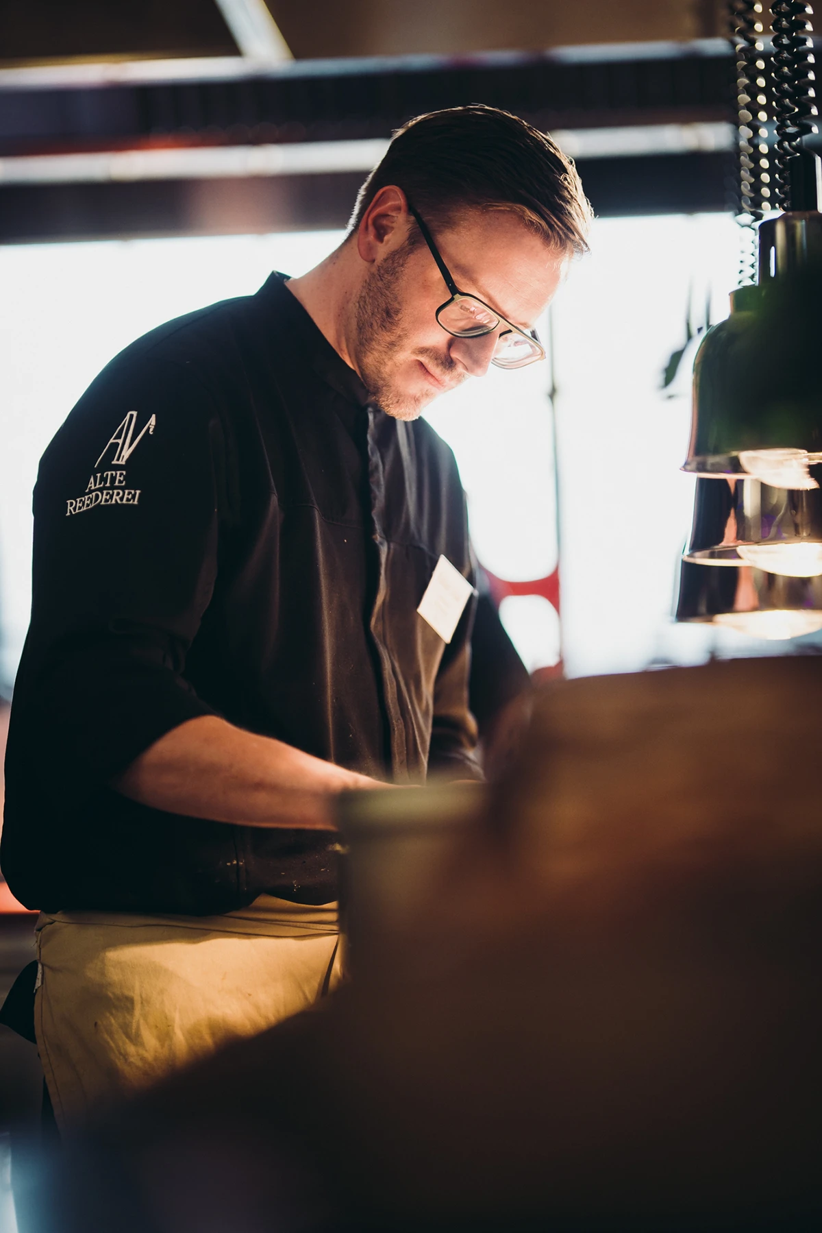 Ein Koch in schwarzer Uniform arbeitet konzentriert in einer Restaurantküche bei warmer Beleuchtung.