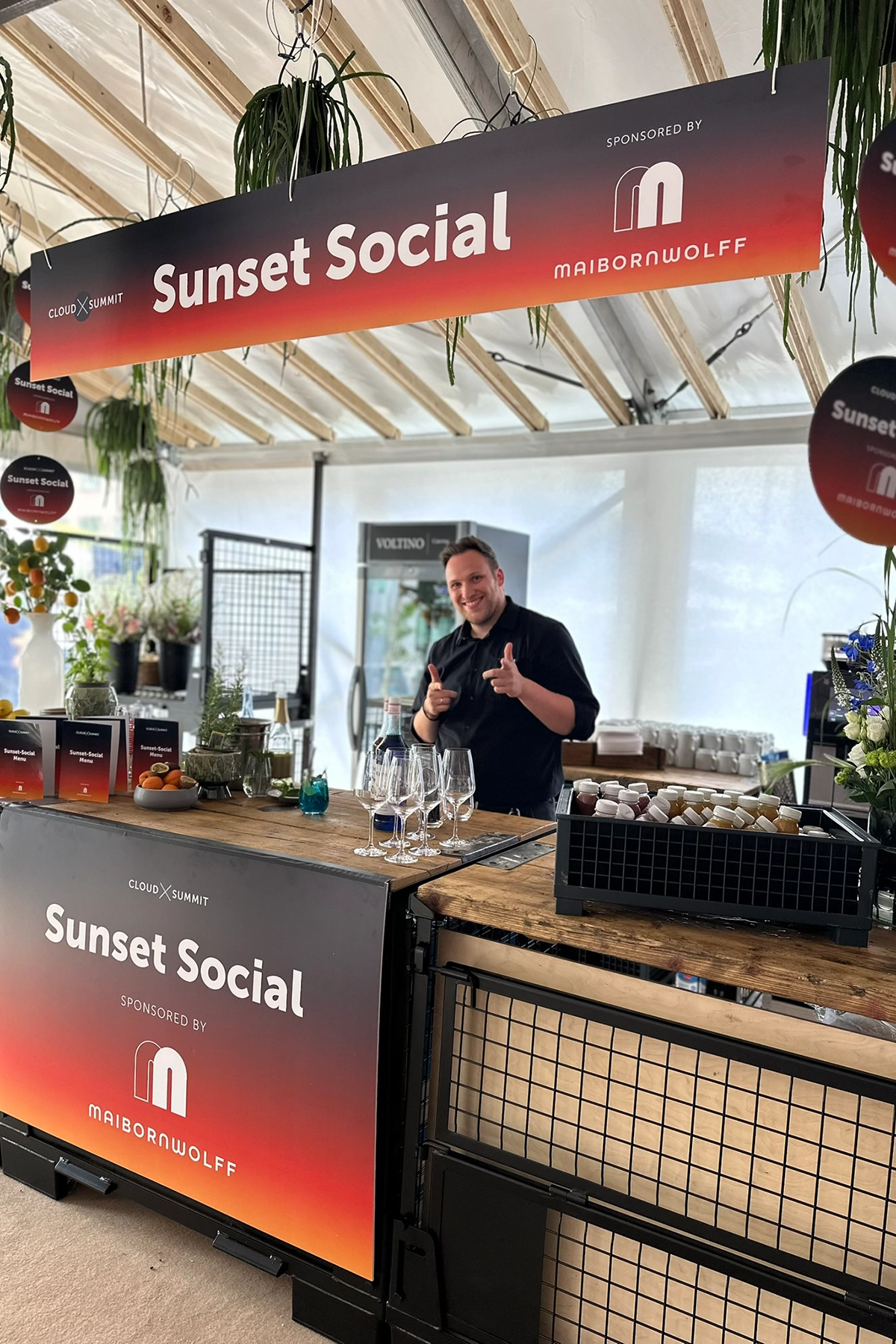 Mann in schwarzer Kleidung hinter Bar mit Gläsern bei Sunset Social-Event in einem dekorierten Zelt.