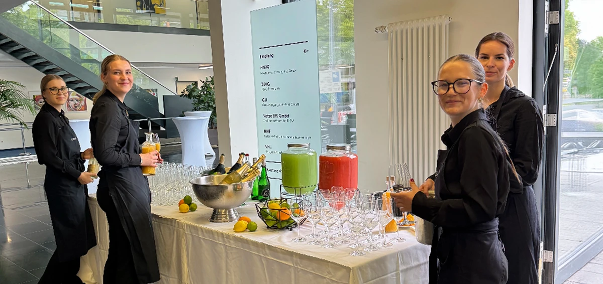 Vier Kellnerinnen in schwarzer Kleidung bedienen ein Getränkebuffet mit Limonaden und Champagner in einem modernen Empfangsbereich.