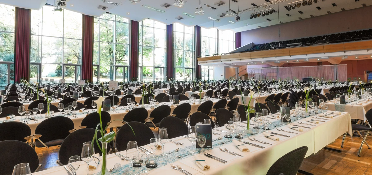 Großer, heller Bankettsaal mit gedeckten Tischen, Gedecken und Blumendekorationen, mit großen Fenstern im Hintergrund.