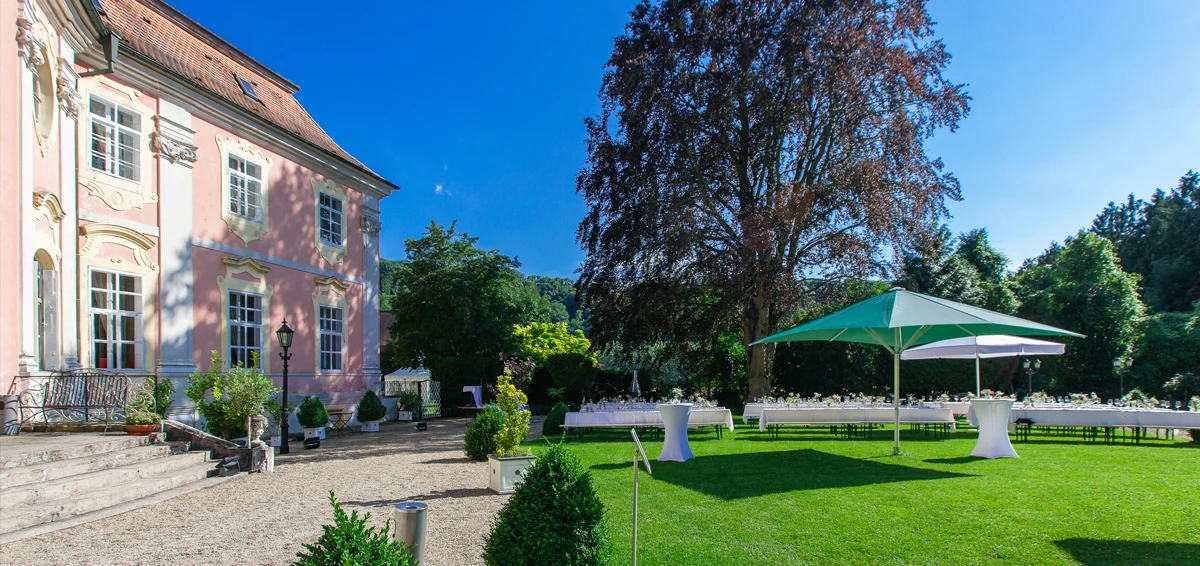 Barockvilla mit Terrasse im Garten, gedeckte Tische und Sonnenschirm im Vordergrund, sonniger Himmel im Hintergrund.