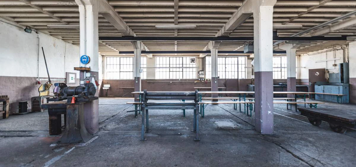 Leere Werkstatt mit Metalltisch in der Mitte, daneben große Fenster, graue Wände und Industriemaschinen im Hintergrund.