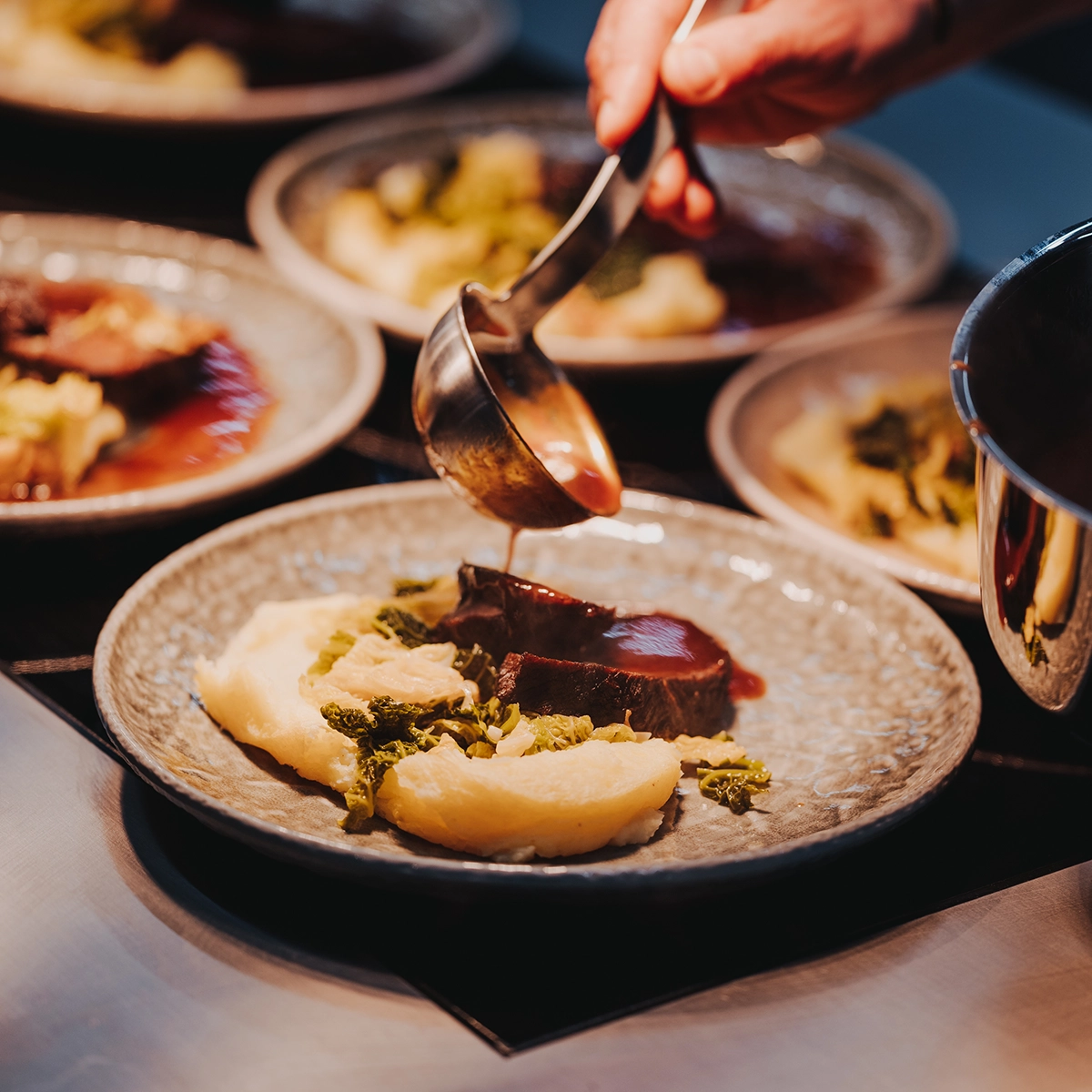 Hand schöpft Soße über Teller mit Fleisch, Kartoffelpüree und Gemüse. Gericht wird angerichtet, warme Farben, Nahaufnahme.