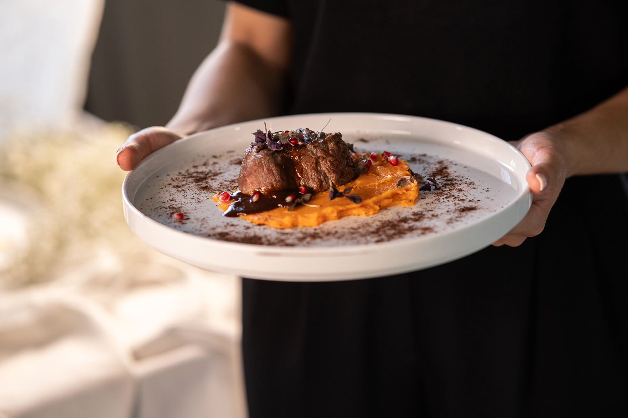 ELEMENTS-Gericht-Feuer Eine Person hält einen Teller mit Rinderfilet auf Süßkartoffelpüree, garniert mit roten Beeren und Kräutern.