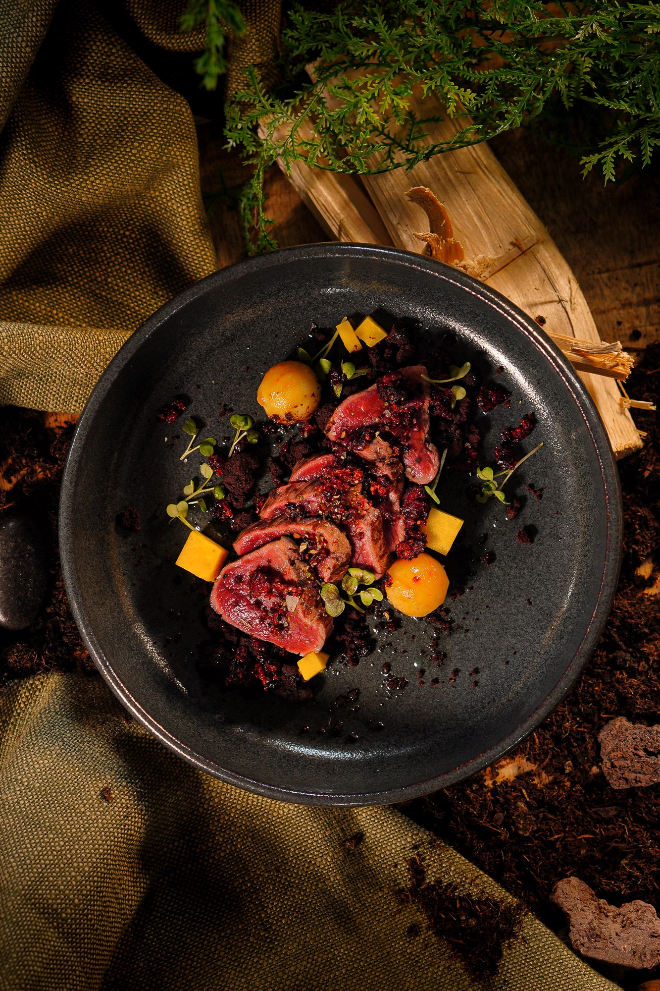 Gourmetgericht mit geschnittenem Fleisch, gelben Würfeln und Tomaten auf schwarzem Teller, garniert mit Kräutern.