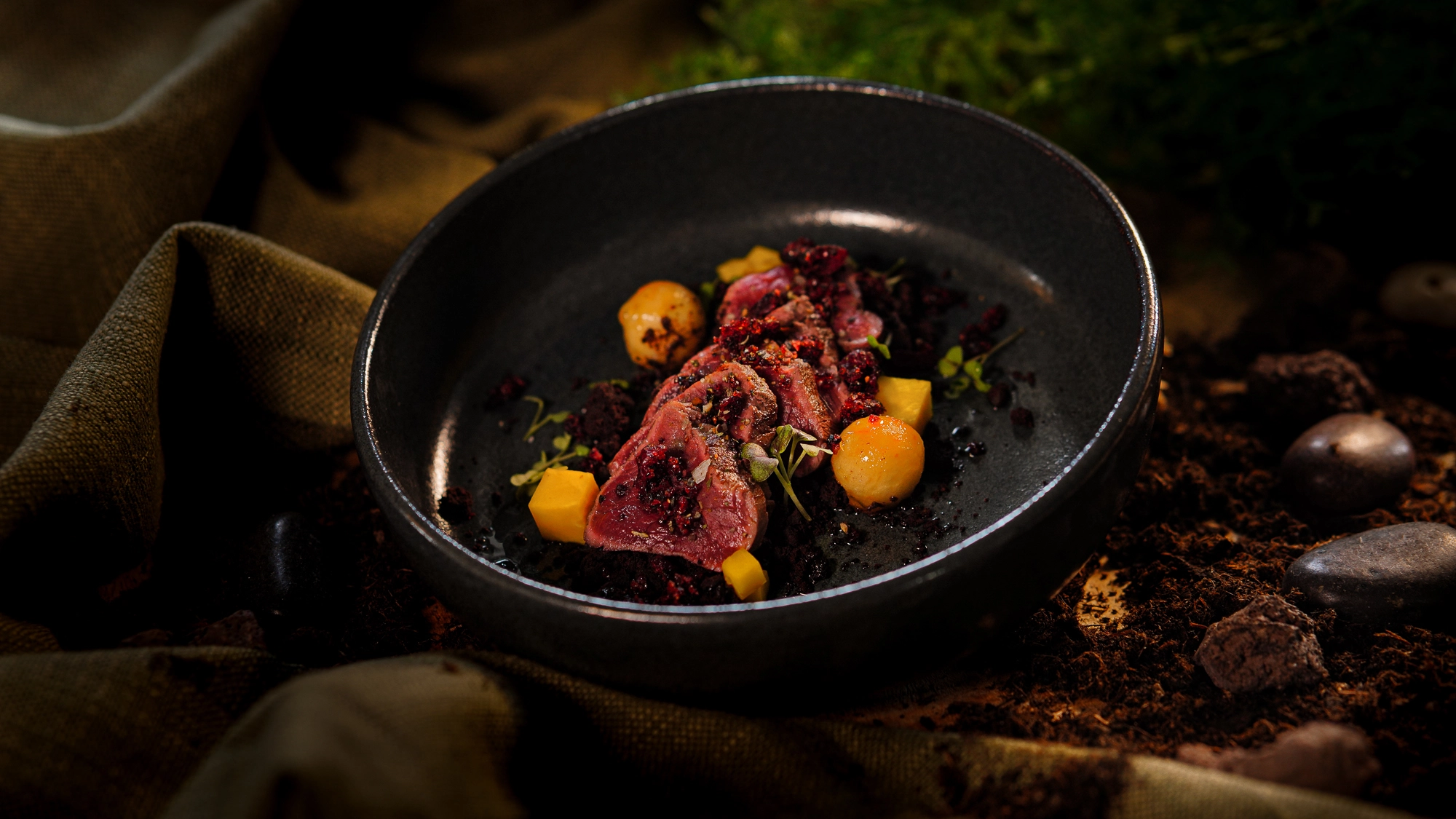 Gourmetgericht in schwarzer Schale: Fleischscheiben mit gelben Kugeln und Kräutern, auf dunklem Tuch und Erddesign serviert.