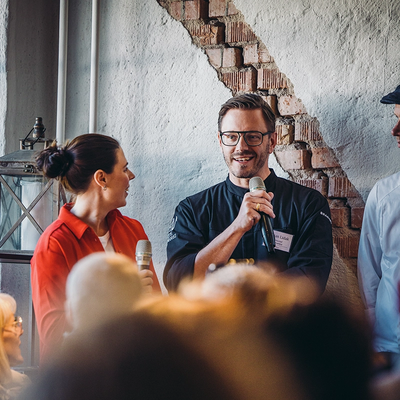 KitchenParty_g-64 Ein Mann und eine Frau halten Mikrofone bei einem Event in einem Raum mit Ziegelwand.