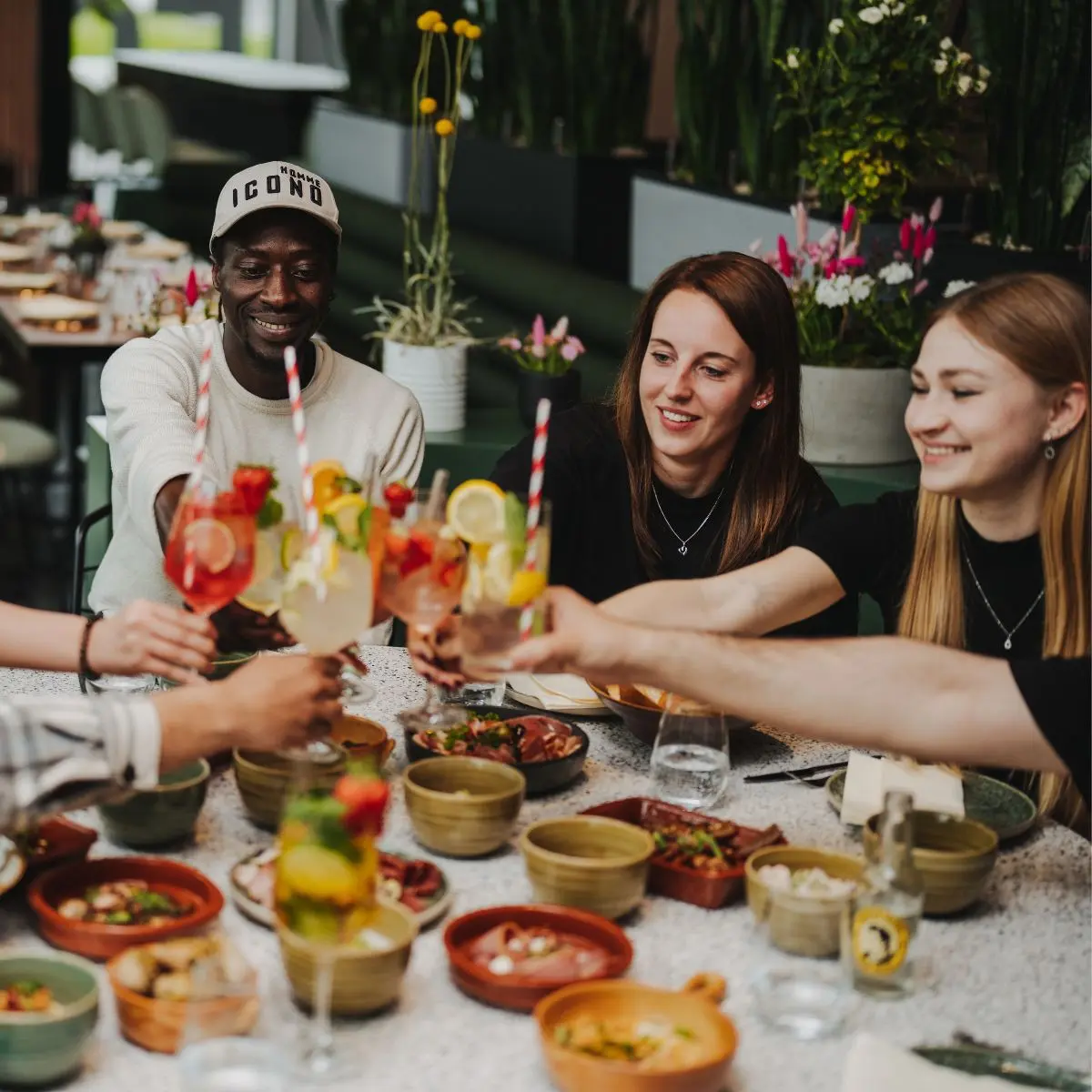 Gruppe von Personen stößt mit bunten Cocktails an einem Tisch voller Schalen und Pflanzen im Hintergrund an.