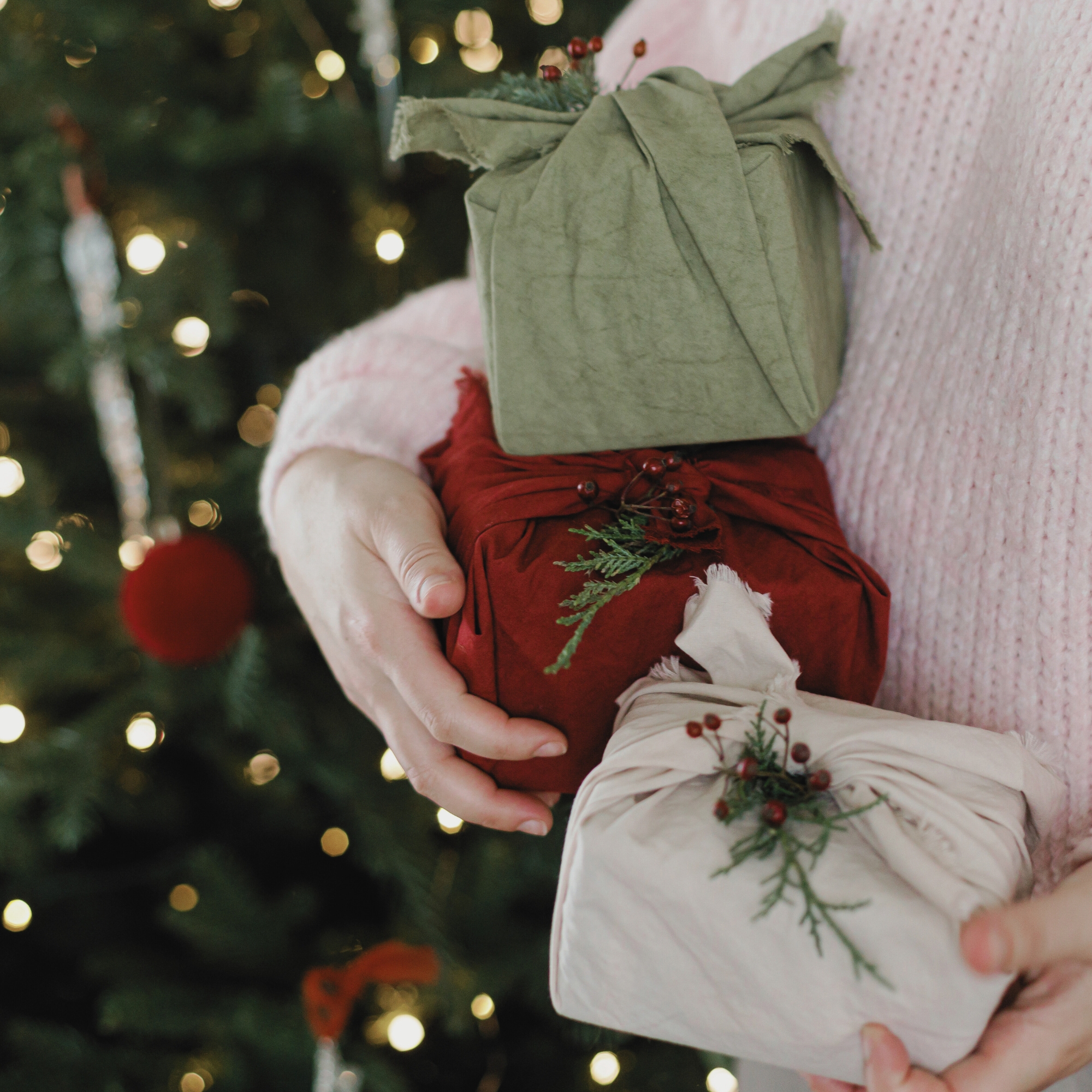 Person hält drei in Stoff eingewickelte Geschenke vor einem festlich geschmückten Weihnachtsbaum.