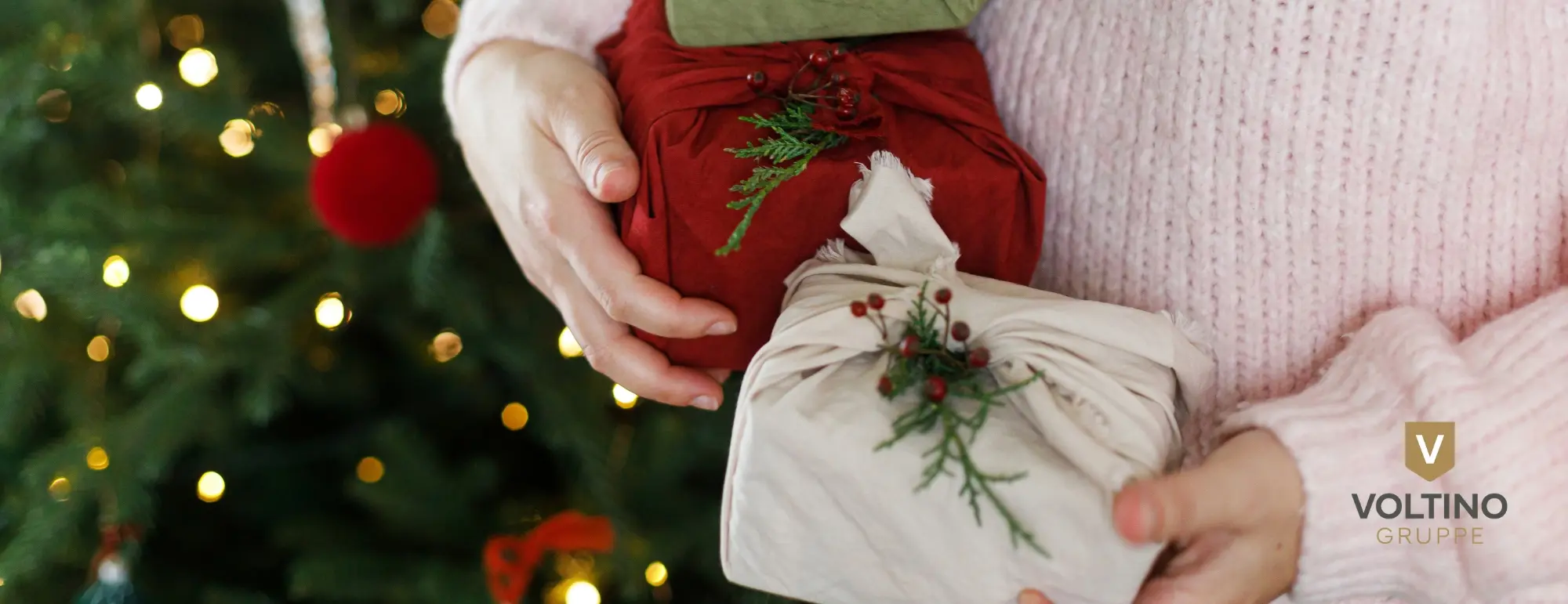 Person hält zwei festlich eingepackte Geschenke vor einem beleuchteten Weihnachtsbaum.
