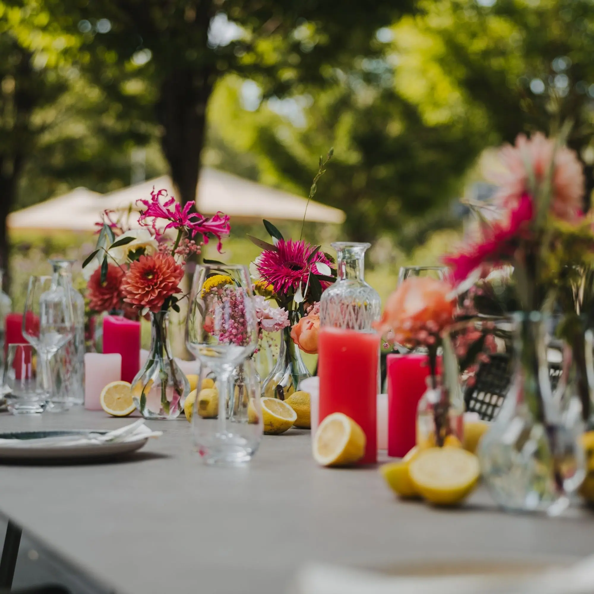 Gedeckter Tisch im Freien mit Blumen, roten Kerzen und Zitronen, umgeben von grüner Natur und Sonnenschirmen.