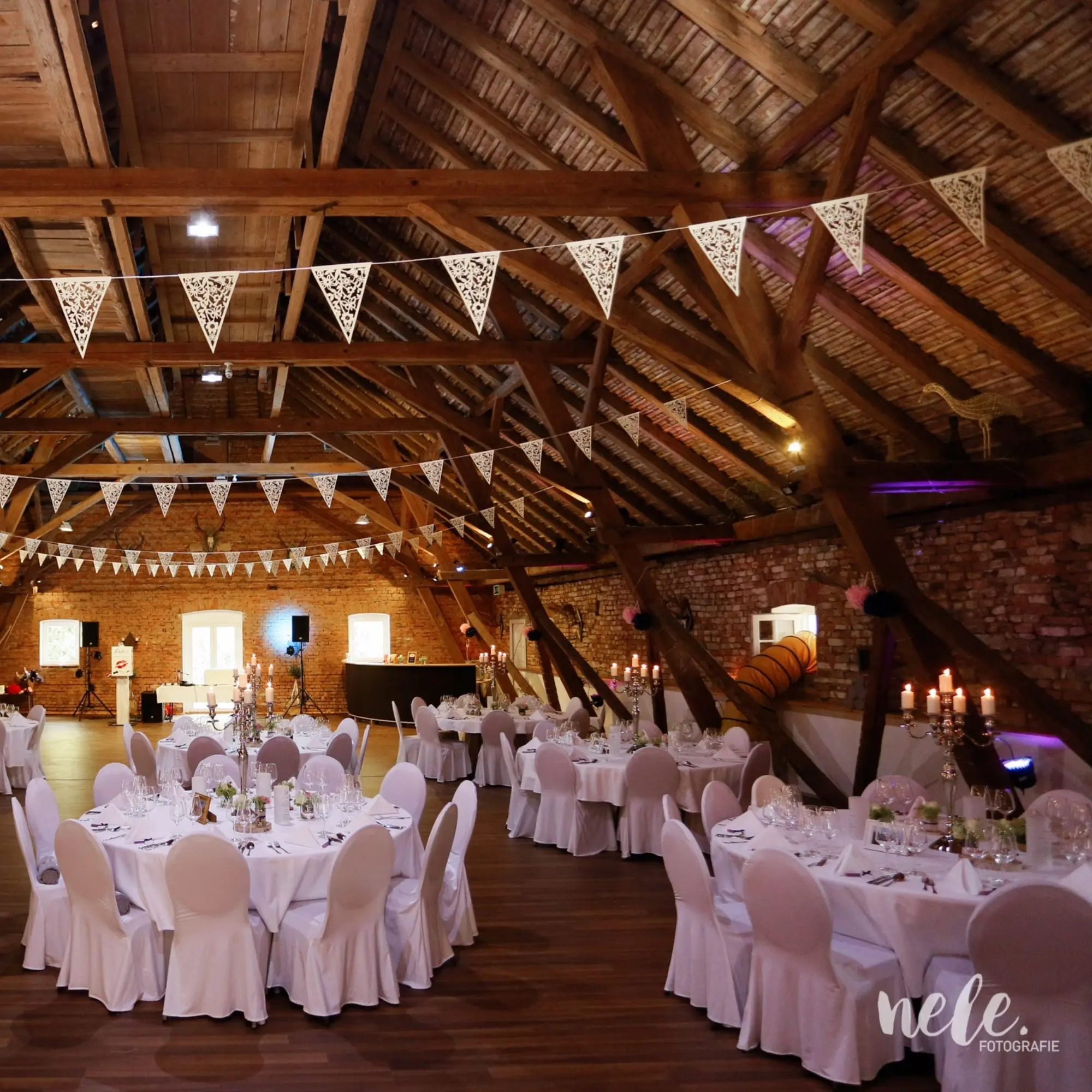 Ein festlich dekorierter Veranstaltungssaal mit Holzbalken, Tischen mit weißen Tischdecken und Girlanden an der Decke.