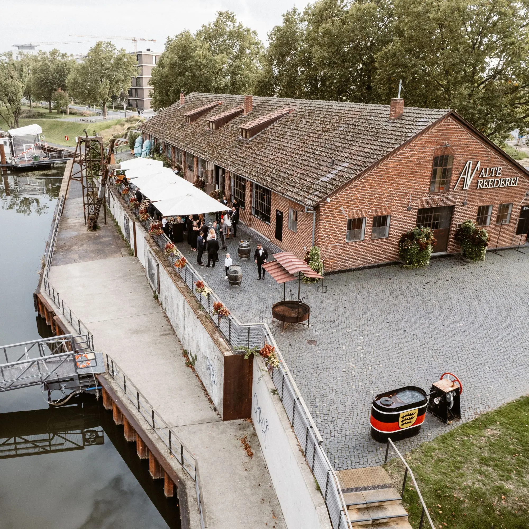 Ehemaliges Backsteingebäude einer Reederei am Wasser, Menschen stehen unter weißen Schirmen auf der Terrasse.