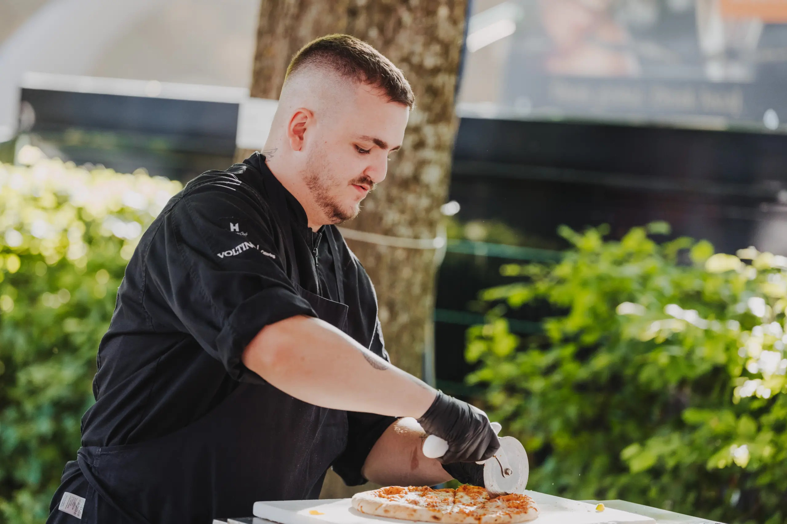 Koch in schwarzer Uniform schneidet Pizza mit einem Pizzaschneider im Freien, umgeben von Bäumen und grüner Kulisse.