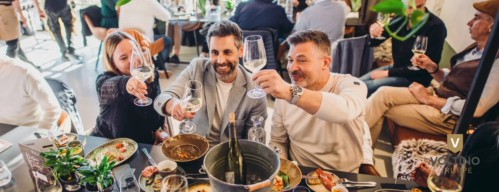 Drei Personen stoßen in einem belebten Restaurant mit Weißweingläsern an, lächelnd und fröhlich.