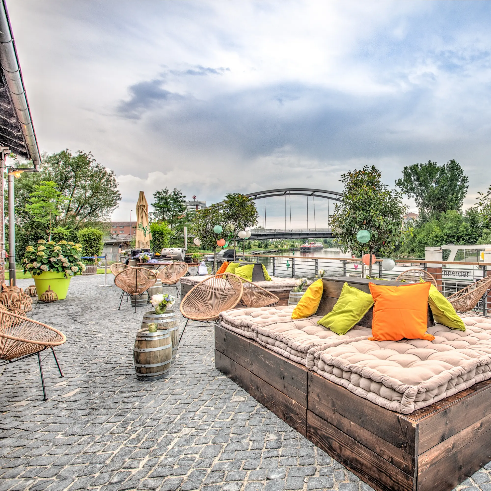 Sonnenterrasse am Fluss mit Sitzgelegenheiten, bunten Kissen und Holzfässern. Brücke und Bäume im Hintergrund.