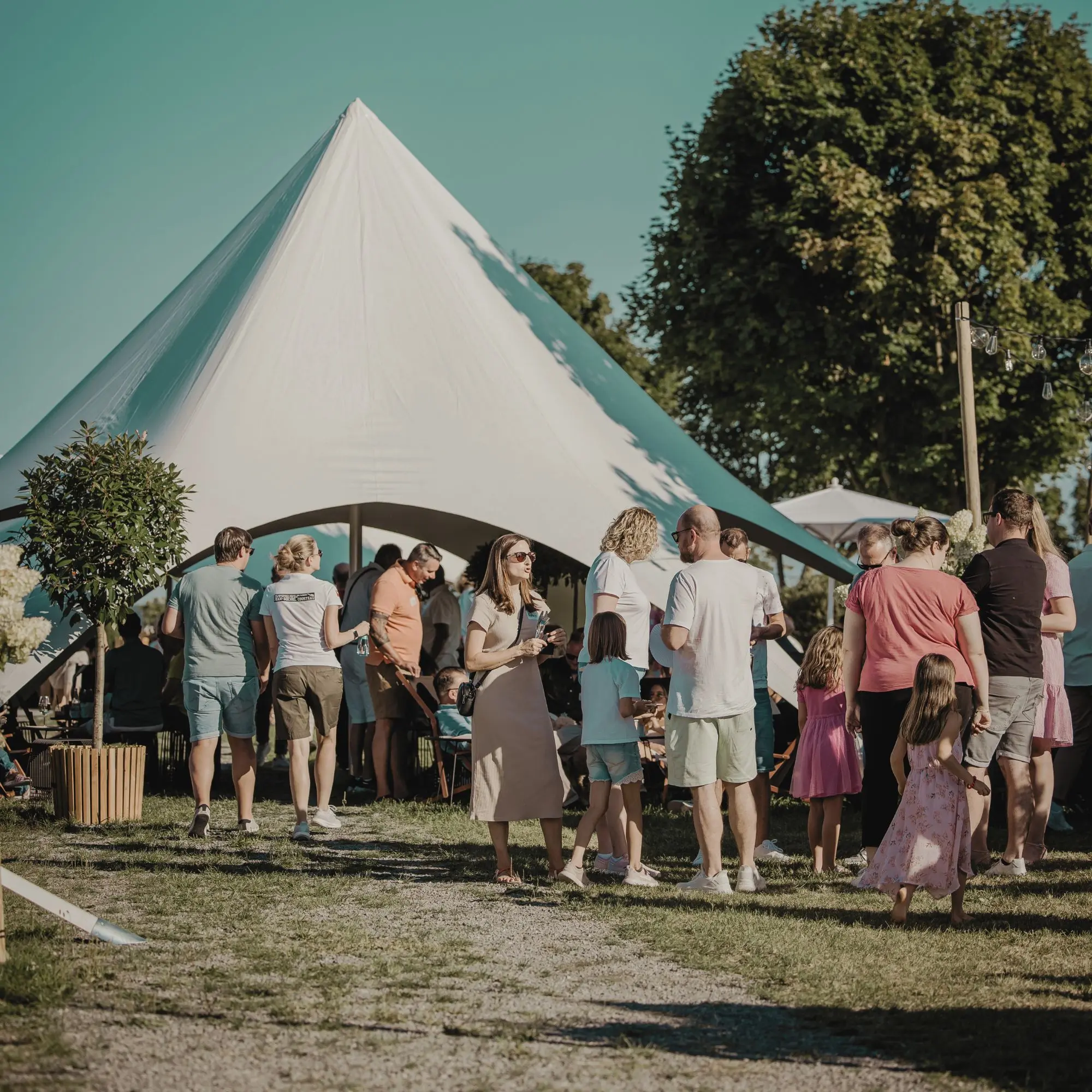 Gruppe von Menschen bei sonnigem Wetter vor einem weißen Festzelt im Freien, Erwachsene und Kinder plaudern.