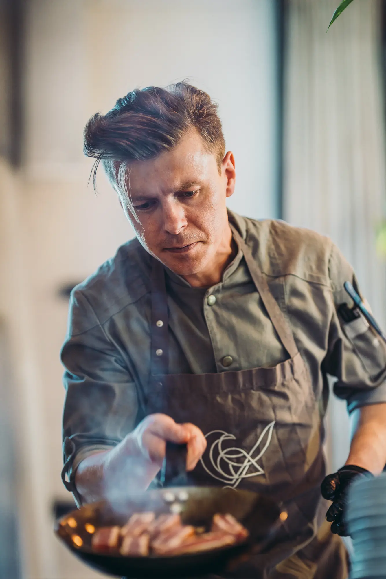Koch in grauer Uniform brät Fleisch in einer Pfanne. Konzentrierter Gesichtsausdruck, Rauch steigt auf.