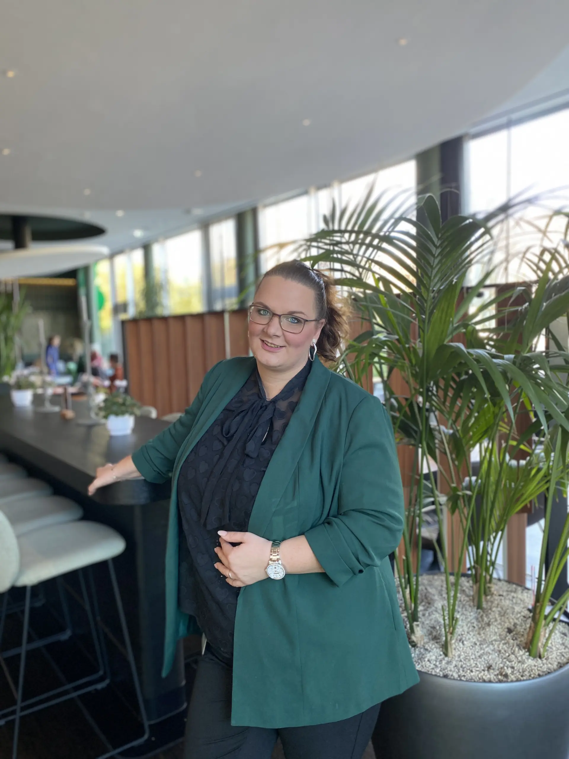 Frau in grünem Blazer steht in modernem Café neben Pflanze, lehnt am Tresen, Tageslicht durch große Fenster.