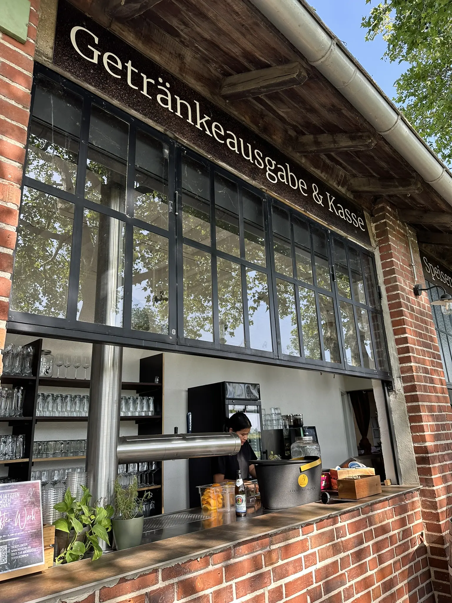 Kiosk-Theke mit Gläsern und Getränken. Eine Person arbeitet an der Getränkeausgabe und Kasse, umgeben von Bäumen.